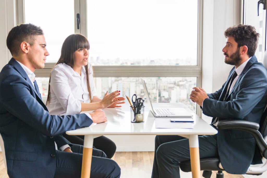 Three professionals in business attire are seated at a desk in a modern office, engaged in a serious discussion, with a city view visible through large windows.