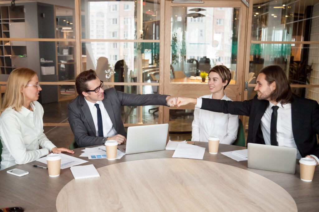 Four colleagues are seated around a conference table in a modern office, with two of them smiling and doing a fist bump, suggesting teamwork and a positive atmosphere.