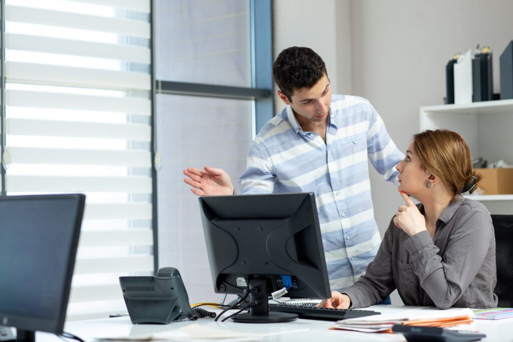 A man is standing and gesturing while speaking to a woman seated at a desk, who appears to be listening thoughtfully in a modern office setting with computers and documents around.